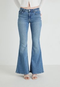 Mujer vestida con jeans acampanados de color azul claro y sandalias blancas de tiras, de pie contra un fondo claro y liso.