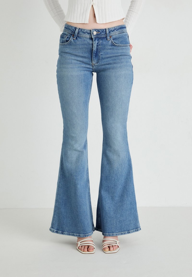 Mujer vestida con jeans acampanados de color azul claro y sandalias blancas de tiras, de pie contra un fondo claro y liso.