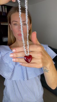 Ketting met een glanzende rode harthanger, geregen aan een delicate ketting van kleine witte parels, tegen een achtergrond van zachte kleuren.