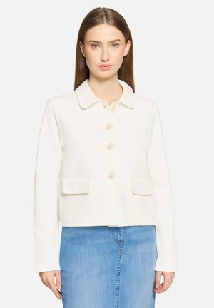 Kobieta o prostych brązowych włosach nosi białą, teksturowaną kurtkę zapinaną na guziki z przednimi kieszeniami oraz jasnoniebieską spódnicę dżinsową.