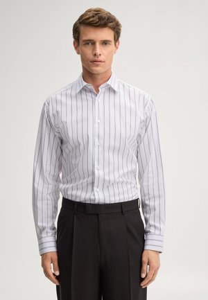 Junger Mann, der ein weißes Hemd mit dünnen schwarzen vertikalen Streifen und eine schwarze Hose trägt, steht vor einem schlichten hellen Hintergrund.
