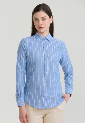 Femme aux cheveux bruns et raides porte une chemise bleu clair à manches longues avec des rayures verticales blanches et un pantalon beige, debout, la main dans la poche.