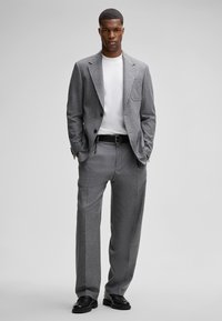 Herrenanzug aus grauem Wollstoff mit tailliertem Schnitt, einreihigem Sakko, Tasche Detail, kombiniert mit einem weißen Rundhalsausschnitt-Shirt und schwarzen Schuhen.