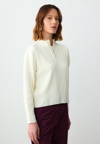 Pull en crème à côtes avec col zippé jusqu'au quart, manches longues et coupe décontractée, associé à un pantalon violet foncé.