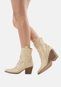 Botas de cuero beige hasta el tobillo con patrones florales en relieve, cierre de cremallera y un tacón de madera apilada. Textura suave y diseño de punta afilada.