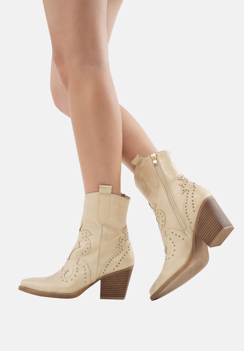 Botas de cuero beige hasta el tobillo con patrones florales en relieve, cierre de cremallera y un tacón de madera apilada. Textura suave y diseño de punta afilada.