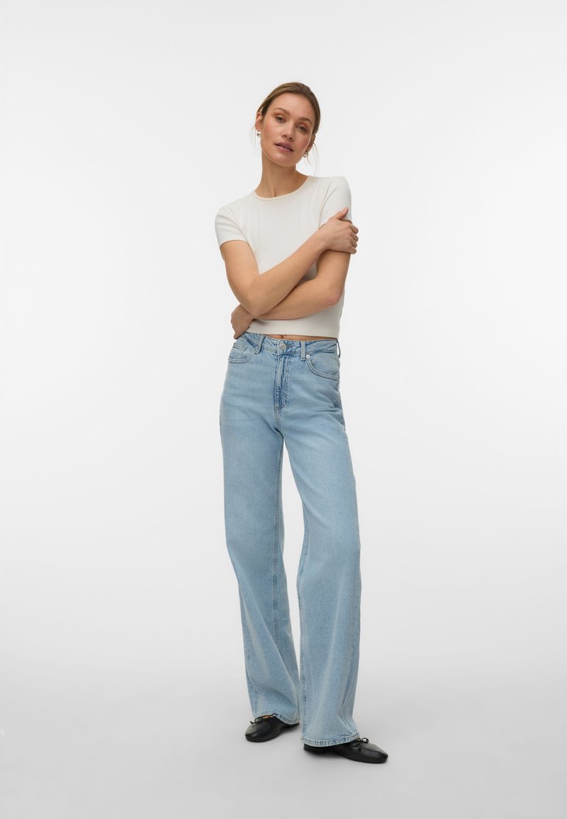 Vrouw die een wit kortärmelig shirt, lichtblauwe wijde spijkerbroek en zwarte platte schoenen draagt, met gekruiste armen staand tegen een witte achtergrond.