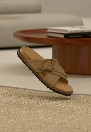 Ταμπά σανδάλι με μεγάλη αγκράφα και σταυρωτά λουράκια πάνω σε υφασμένο μπεζ χαλί, δίπλα σε στρογγυλό ξύλινο τραπεζάκι με βιβλία.