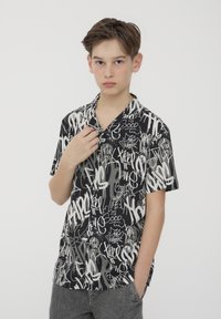 Camicia a maniche corte con bottoni in nero e grigio, decorata con una stampa in stile graffiti. Il tessuto sembra leggero, con una vestibilità comoda e colletto da campeggio.