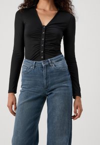 Black long-sleeve bodysuit with a V-neck and gathered front, featuring a row of black buttons. Paired with light blue high-waisted jeans.