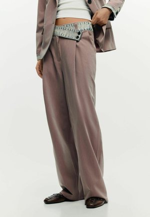 Person trägt breite, mauvefarbene Hosen mit überlappendem Denim-Bund, ein weißes Crop-Top, einen mauvefarbenen Blazer und braune Schuhe.