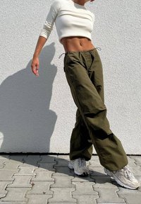 Bílý nákrutový crop top, olivově zelené široké kalhoty s šňůrkou a světle šedé tenisky. Texturovaná zeď v pozadí.