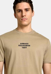 Beige bomulds t-shirt med "ARMANI EXCHANGE 1991" broderet i sort, rund hals, korte ærmer og et tætsiddende design.