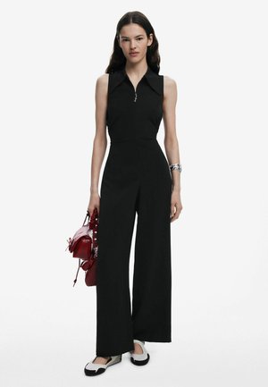 Black sleeveless jumpsuit with a collar and zipper detail, wide-leg design. Paired with a small red handbag and black-and-white footwear.