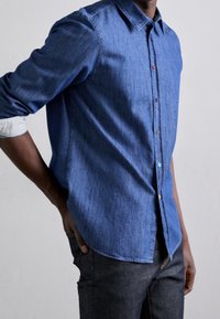 Sininen farkkupaita, jossa on napillinen kaulus, moniväriset napit ja käännetyt raidalliset hihat. Päällä tummat farkut.