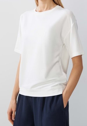 Bílá krátkosrstá košile s kulatým výstřihem, vyrobená z hladké tkaniny; má uvolněný střih a jemný detail na bočním švu.