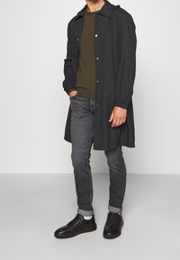 Schwarze leichte Jacke mit Druckknöpfen, langen Ärmeln und lockerer Passform, getragen über einem dunkelgrünen Shirt und grauen Jeans. Schwarze Sneakers runden den Look ab.