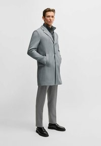 Grå ullblandad kappa med snedskurna slag och två fickor, lager på lager över en svart jacka; i kombination med grå randiga byxor och svarta skor.