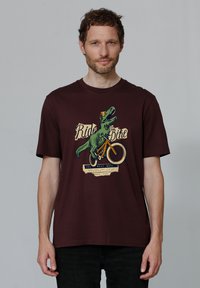 Bordeaux katoenen t-shirt met een dinosaurustekening die op een fiets rijdt, met de tekst "Fiets Rijden" en "de beste dagen." Korte mouwen, ronde hals.