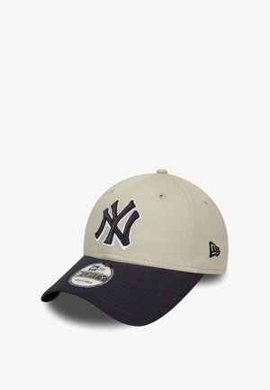 Beige Baseballkappe mit einem dunkelblauen Schirm, mit einem schwarzen und weißen gestickten NY-Logo auf der Vorderseite und einem verstellbaren Riemen auf der Rückseite.