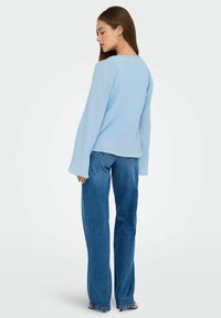 Top azzurro chiaro a maniche lunghe in tessuto testurizzato con vestibilità rilassata, abbinato a jeans a gamba larga blu scuro e orecchini semplici.