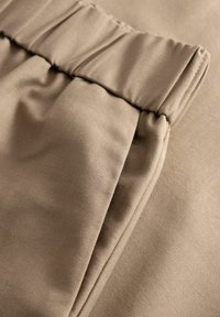 Lähikuva beigen värisistä kangashousuista, joissa on kuminauhavyötärö ja näkyvät saumat sivutaskuissa.