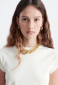 Weißes, eng anliegendes T-Shirt mit kurzen Ärmeln, kombiniert mit mehreren goldenen Kettenhalsreifen und strukturierten goldenen Creolen. Glattes Stoffmaterial.