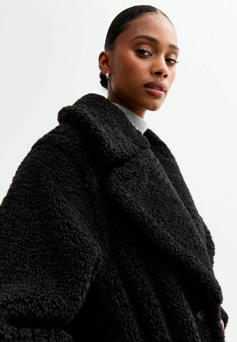 Czarny oversize płaszcz wykonany z pluszowego materiału, z dużym kołnierzem i teksturowaną powierzchnią. Projekt obejmuje guziki oraz luźny krój.