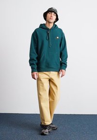 Zeleni flisani dukserica s kenguruh džepom, bež hlače i sive tenisice. Outfit se odlikuje minimalističkim dizajnom i opuštenim krojem.