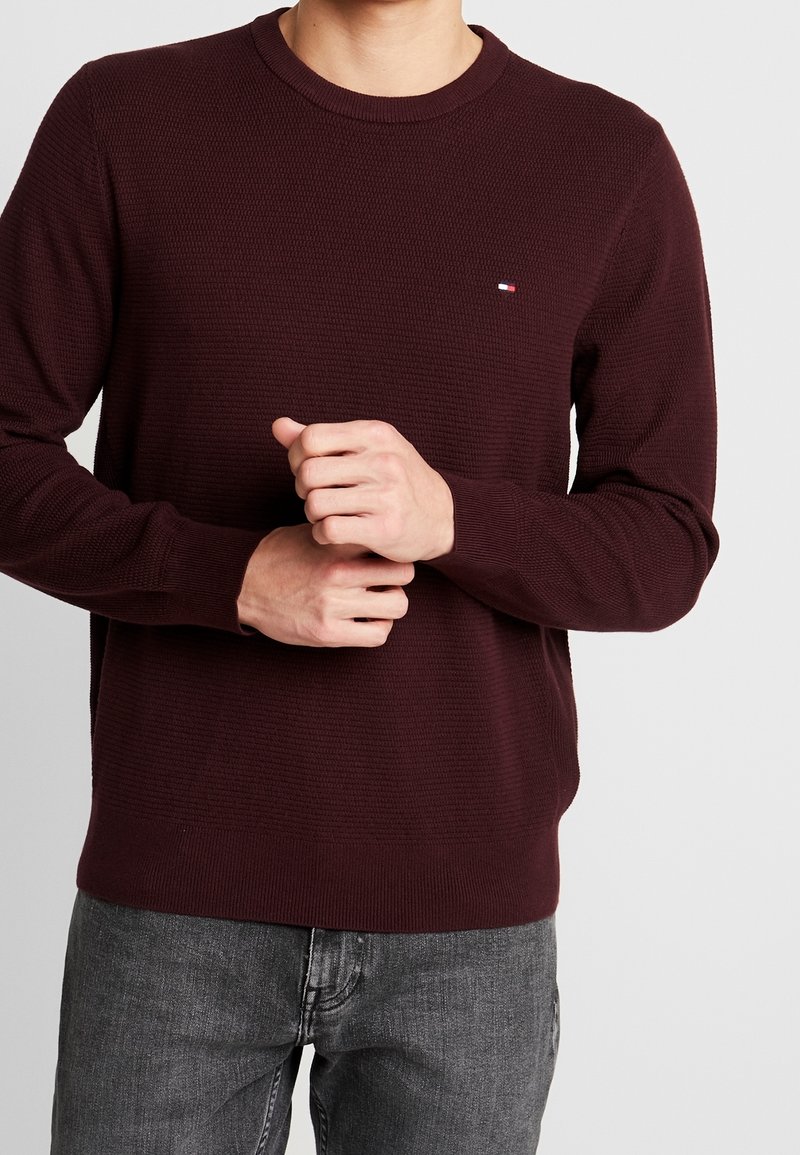 Homme portant un pull texturé bordeaux foncé avec un petit logo rouge, blanc et bleu sur la poitrine, associé à un jean gris.