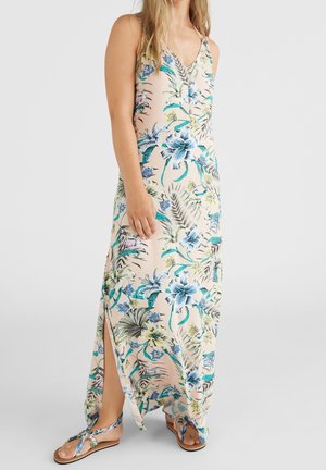 Kvinde iført en ærmeløs, blomsterprintet maxikjole med sidesprække og matchende blomsterprintede sandaler mod en ensfarvet baggrund.