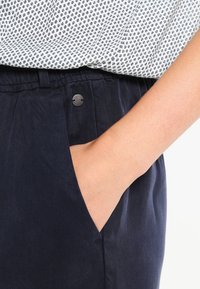 Námořnické modré kalhoty s texturovanou tkaninou, které mají boční kapsu a kovovou sponu na pase, v kombinaci se vzorovaným bílým topem.