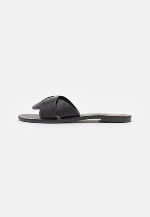 Sort flad sandal med brede krydsede remme og åben tå, vist i profil mod en hvid baggrund.