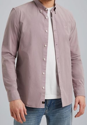 Mand iført en åbnet lys mauve langærmet skjorte over en hvid t-shirt og blå jeans, stående foran en ensfarvet baggrund.