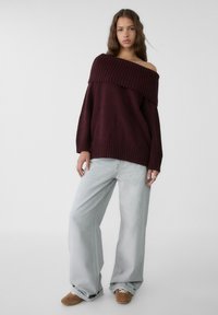 Burgundska off-shoulder puli z rebrasto teksturo, v kombinaciji z ohlapnimi svetlo modrimi kavbojkami in rjavimi natikači. Preprost, sproščen kroj.