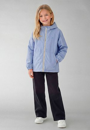 Fată tânără zâmbitoare, purtând o jachetă cu glugă albastru deschis cu fermoar, pantaloni negri cu țesătură largă și adidași albi, stând pe un fundal simplu.