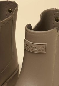 Tan-nahkaiset nilkkurit, joissa on teksturoitu pinta ja sivussa bränditekstiilipatchi. Pyöreä kärki ja matala profiili.