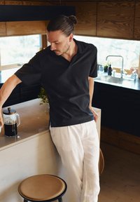 Zwart geribbeld poloshirt met een relaxte pasvorm, gecombineerd met witte linnen broek. Een Franse pers staat op een aanrecht in een moderne keuken setting.
