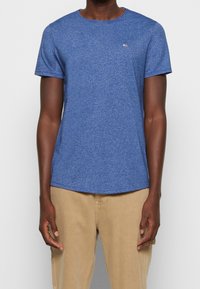 Camiseta corta de manga azul con cuello redondo, tejido texturizado y un pequeño logo de marca en la parte superior izquierda. Combinada con pantalones beige.