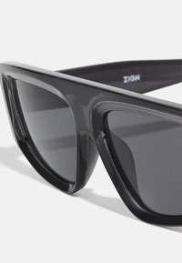 Gafas de sol de plástico negro con un diseño afilado y angular. Presentan lentes oscuros y una textura suave. El nombre de la marca "ZIGN" es visible en la parte interior de la patilla.