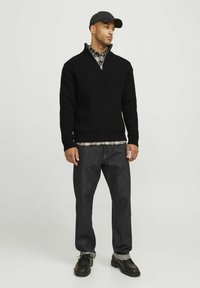 Schwarzer Strickpullover mit Viertel-Reißverschluss, getragen über einem karierten Flanellhemd. Kombiniert mit dunklen Jeans und schwarzen Schnürschuhen. Lässiges Outfit.