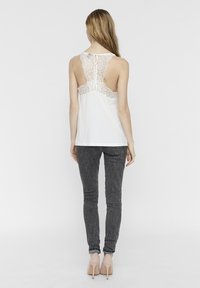 Kvinna med långt hår iförd en vit linne med spets på ryggen, mörkgrå skinny jeans och beige högklackade skor, stående vänd bort.