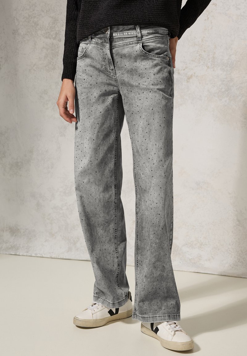 Grijze denim jeans met een relaxte pasvorm, voorzien van lichte slijtage en kleine studdetails. Draagt met witte sneakers.