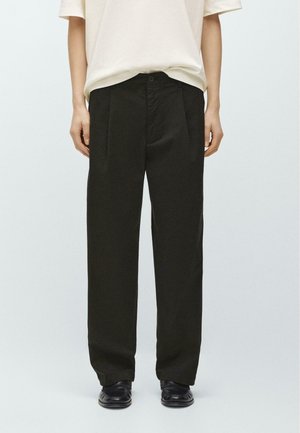 Persona che indossa pantaloni neri ampi, scarpe nere e una camicia beige chiaro a maniche corte, in piedi davanti a uno sfondo chiaro e semplice.