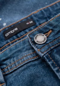 Blå denimtyg med orange sömmar, med en silvrig knapp märkt "Ombre Denim" och en svart etikett som står "H32." Strukturerad yta med lätt blekning.