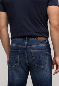 Mężczyzna w ciemnoniebieskich dżinsach z pomarańczowymi przeszyciami i granatowej koszulce, przedstawiony od talii w dół i z tyłu.