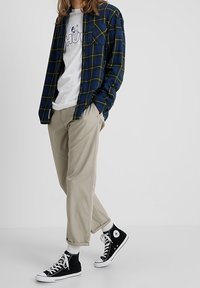 Camicia di flanella a quadri blu e verde sovrapposta a una maglietta grafica bianca, abbinata a pantaloni beige e sneaker neri alte. Calzini bianchi visibili.