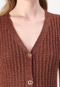 Rostfarbener Cardigan mit V-Ausschnitt, geripptem Muster und goldfarbenen Knöpfen mit kreisförmigem Design. Strickmaterial mit Schimmer.