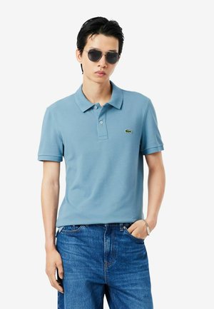 Giovane uomo con occhiali da sole neri, polo celeste con logo coccodrillo e jeans blu, in piedi con una mano in tasca.