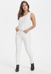 Bijela majica bez rukava opuštenog kroja, uparena s uskim bijelim trapericama. Crne gležnjače s metalnim detaljima upotpunjuju look.